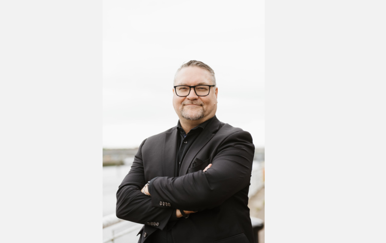 Dr. Jason R. Frank standing outdoors with arms crossed near a waterfront.